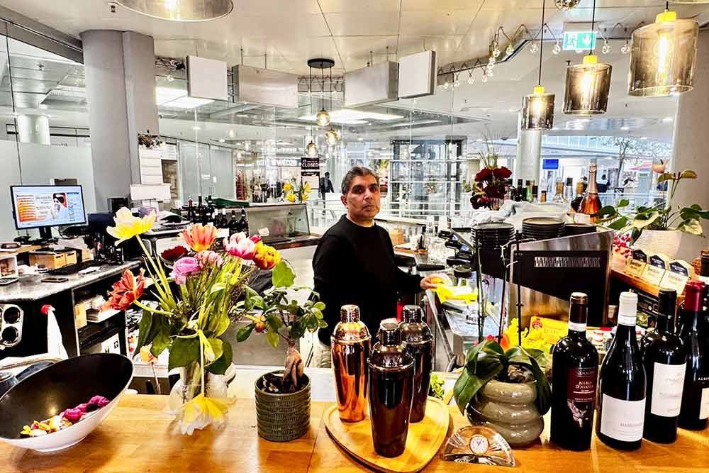 Ein Barkeeper steht hinter der Bar, im Vordergrund sind Blumen und Flaschen zu sehen. Hell beleuchteter Ladeninnenraum.