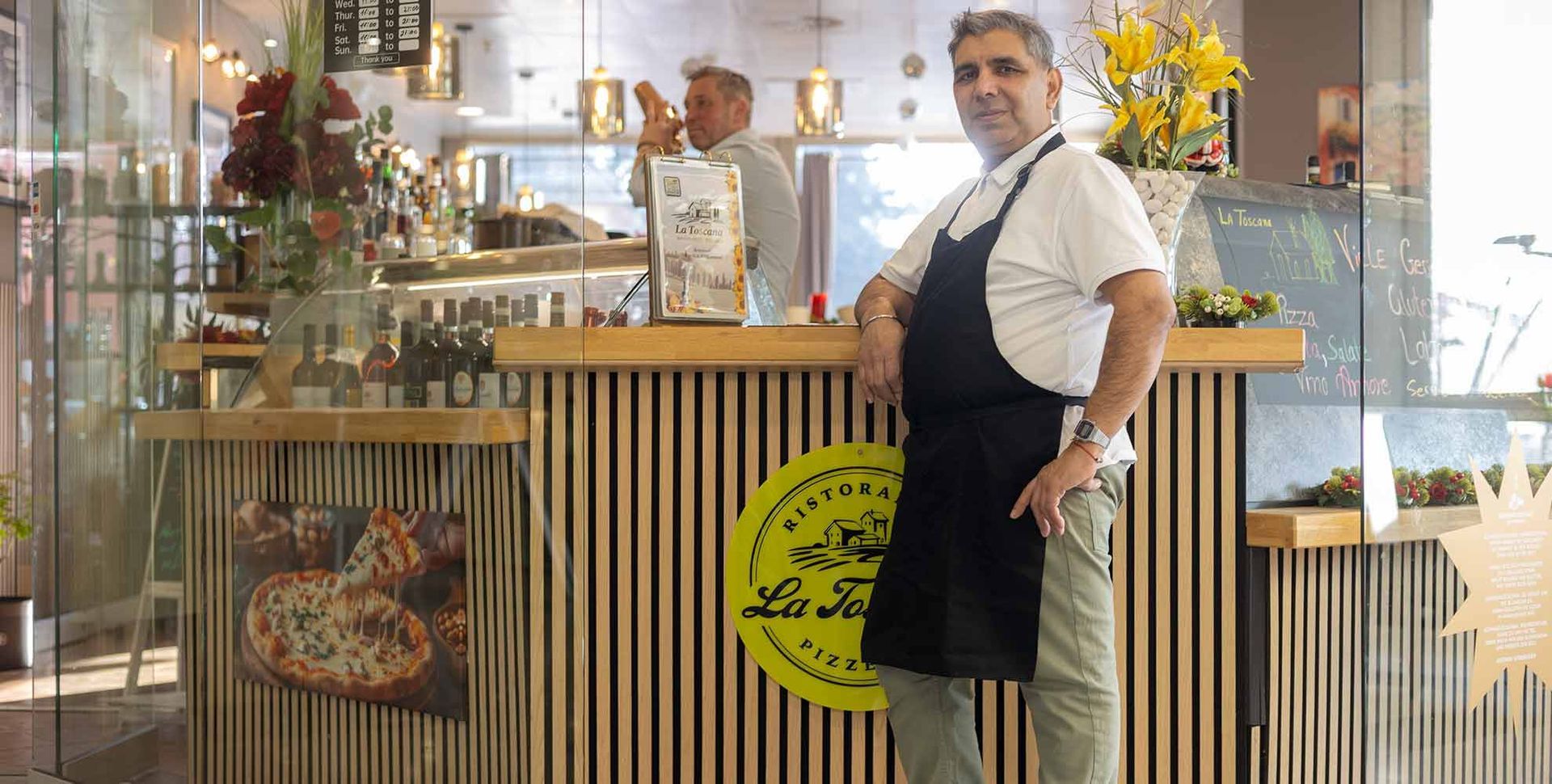 Ein Mann mit Schürze steht an einem Restauranttresen. Im Hintergrund ist eine weitere Person zu sehen.