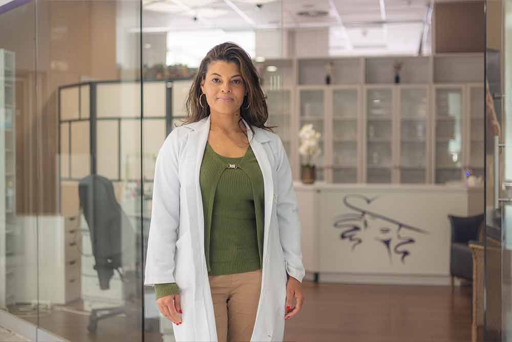 Eine Frau im Laborkittel steht im Eingang einer Klinik. Olivgrüner Pullover, beigefarbene Hose, heller Hintergrund.