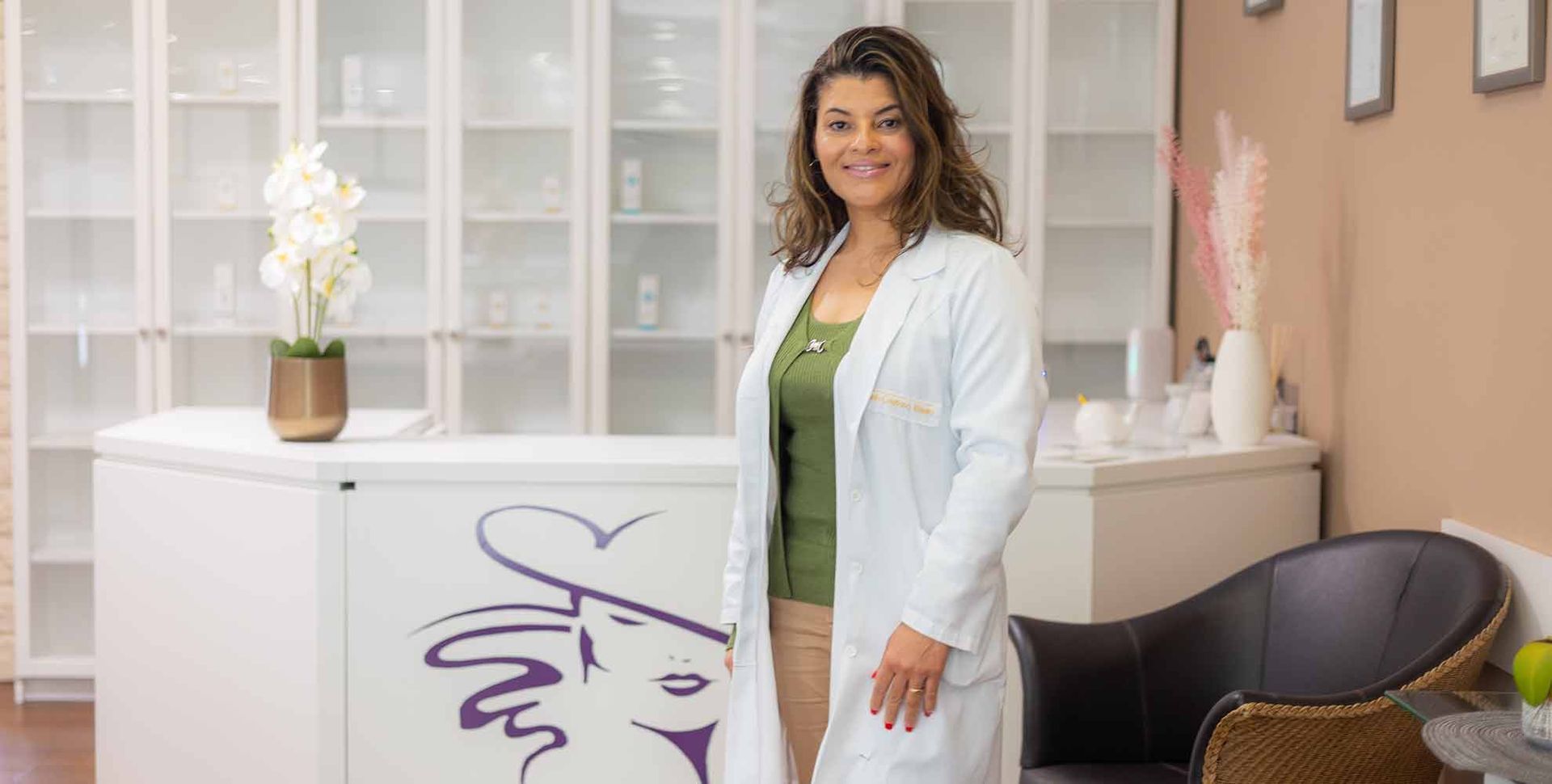 Eine Frau im Laborkittel steht in einer Klinik vor einem Empfangstresen und Regalen.