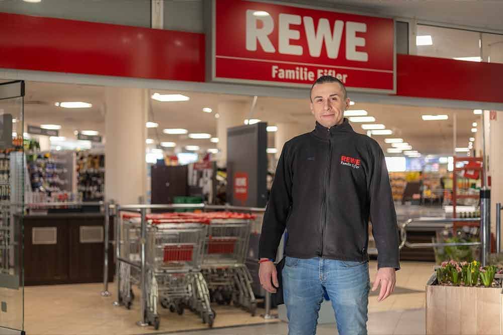 Ein Mann steht vor dem Eingang eines REWE-Supermarkts. Er trägt eine schwarze Jacke und Jeans. Der Laden hat rote Schilder.