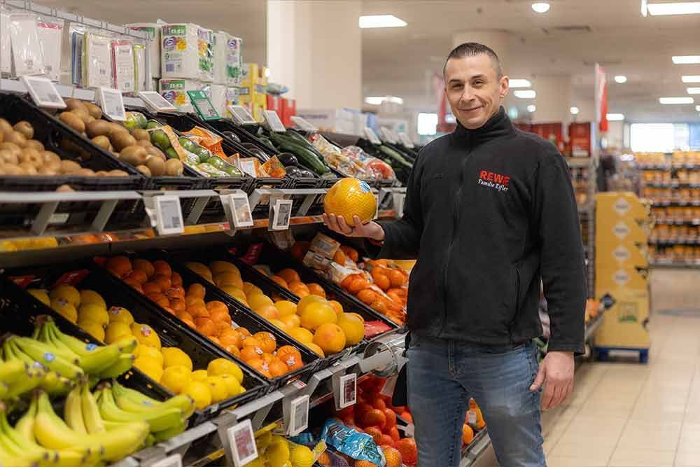 Ein Mann in einer schwarzen Jacke hält eine gelbe Frucht in der Obst- und Gemüseabteilung eines Supermarktes.