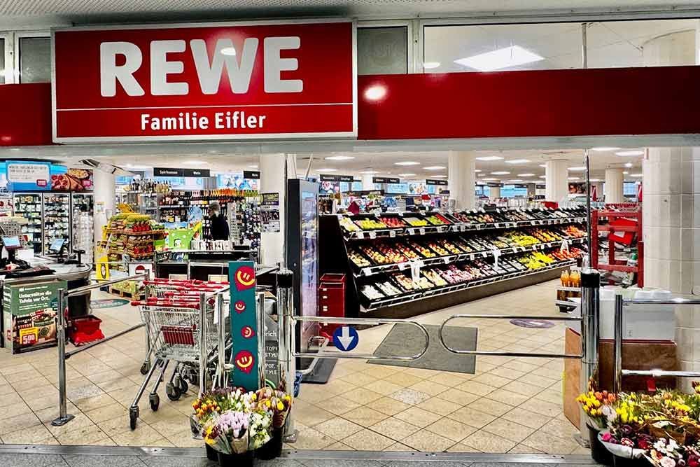 Eingang zum REWE-Supermarkt mit roter Beschilderung und Reihen von Obst- und Gemüseregalen. Einkaufswagen und automatische Türen.