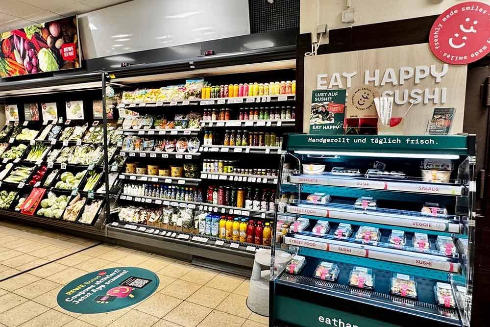 Obst- und Gemüseabteilung sowie Sushi-Auslage in einem Supermarkt. Grünes, frisches Obst und Gemüse, abgefüllte Getränke und zubereitetes Sushi sind zu sehen.