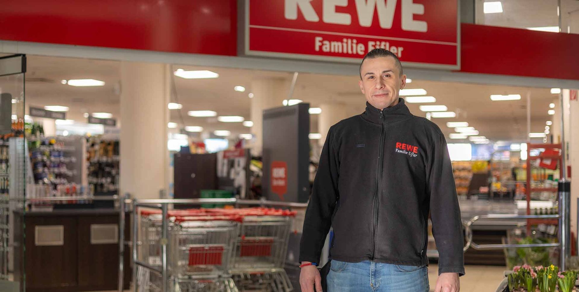 Ein Mann in schwarzer Jacke steht mit Einkaufswagen am Eingang eines REWE-Supermarkts.