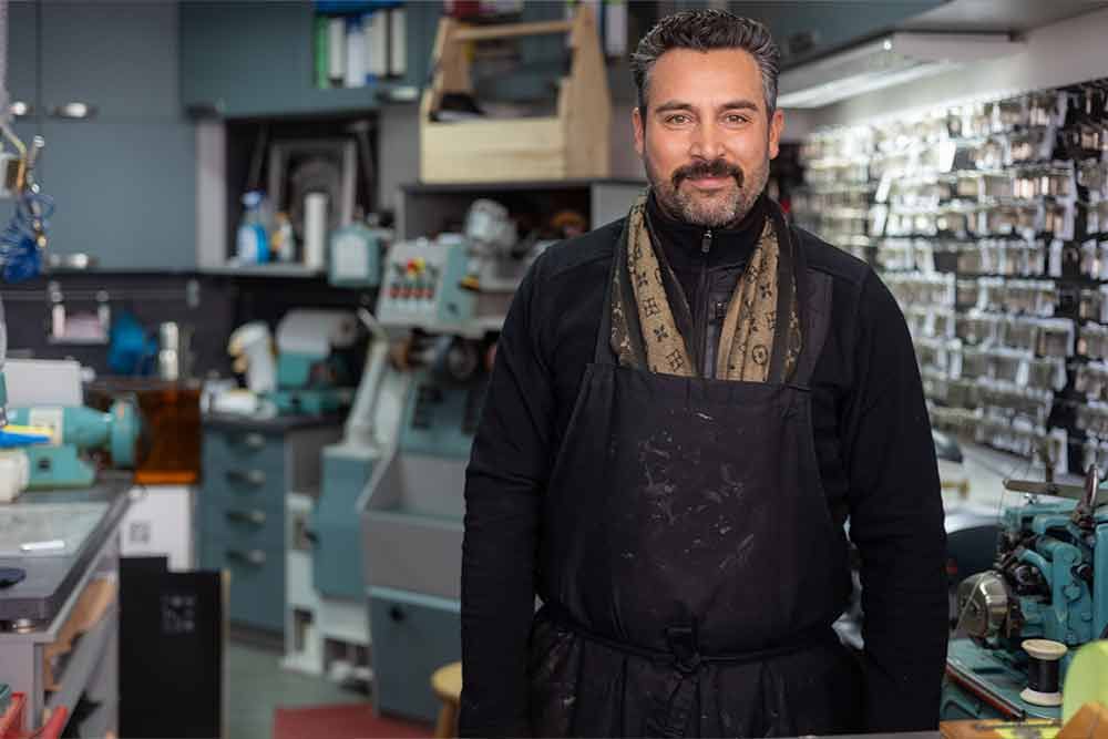 Schuster in der Werkstatt, lächelnd, mit Schürze und Schal bekleidet, Werkzeuge und Ausrüstung im Hintergrund.