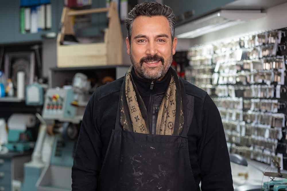 Ein Mann in Schürze lächelt in einem Schlüsseldienstgeschäft mit Werkzeugen zur Schlüsselherstellung und Schlüsselauslagen.