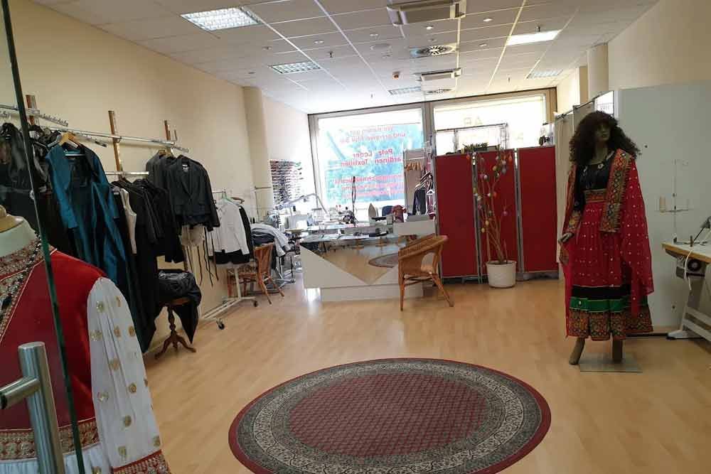 Innenansicht eines Bekleidungsgeschäfts mit Kleiderständern, Schaufensterpuppen, Nähstation und einem Teppich.