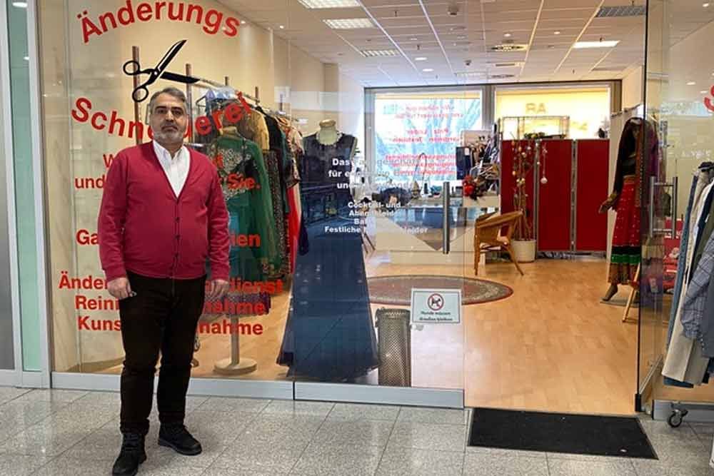 Ein Mann steht vor einem Änderungsatelier. Im Schaufenster sind Kleidungsstücke ausgestellt. Er trägt eine rote Strickjacke und eine braune Hose.