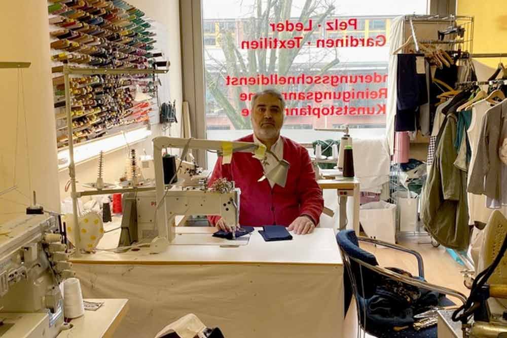 Mann an einer Nähmaschine in einer Werkstatt mit Stoff, Garnrollen und einem Schaufensterschild.