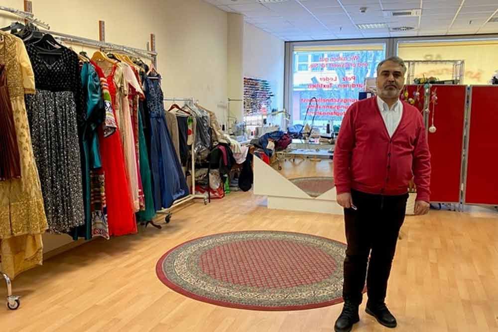 Ein Mann in einem roten Pullover steht in einem Bekleidungsgeschäft, Kleider hängen an Kleiderständern, Teppich liegt auf dem Boden.