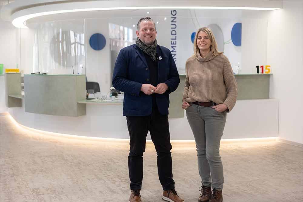 Ein Mann und eine Frau stehen an einem Empfangstresen. Der Mann trägt eine blaue Jacke, die Frau einen Pullover. Helle Büroumgebung.