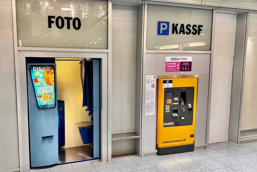 Fotokabine und Parkscheinautomat in einem hell erleuchteten Innenraum. Schilder mit der Aufschrift „Foto“ und „Kasse“.