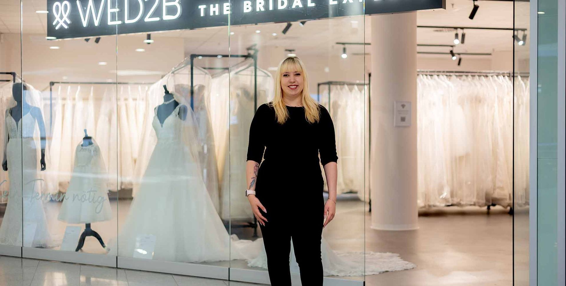 Eine Frau steht vor einem Brautkleidgeschäft und zeigt auf die Auslage.