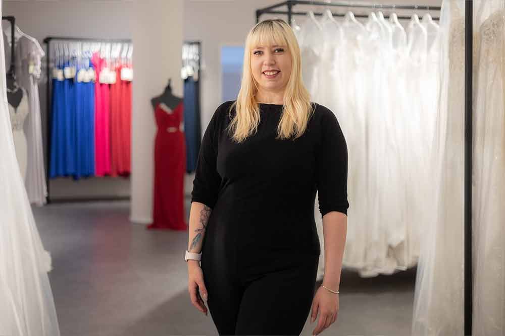 Frau in schwarzem Outfit lächelt in einem Brautmodengeschäft, Kleider im Hintergrund.