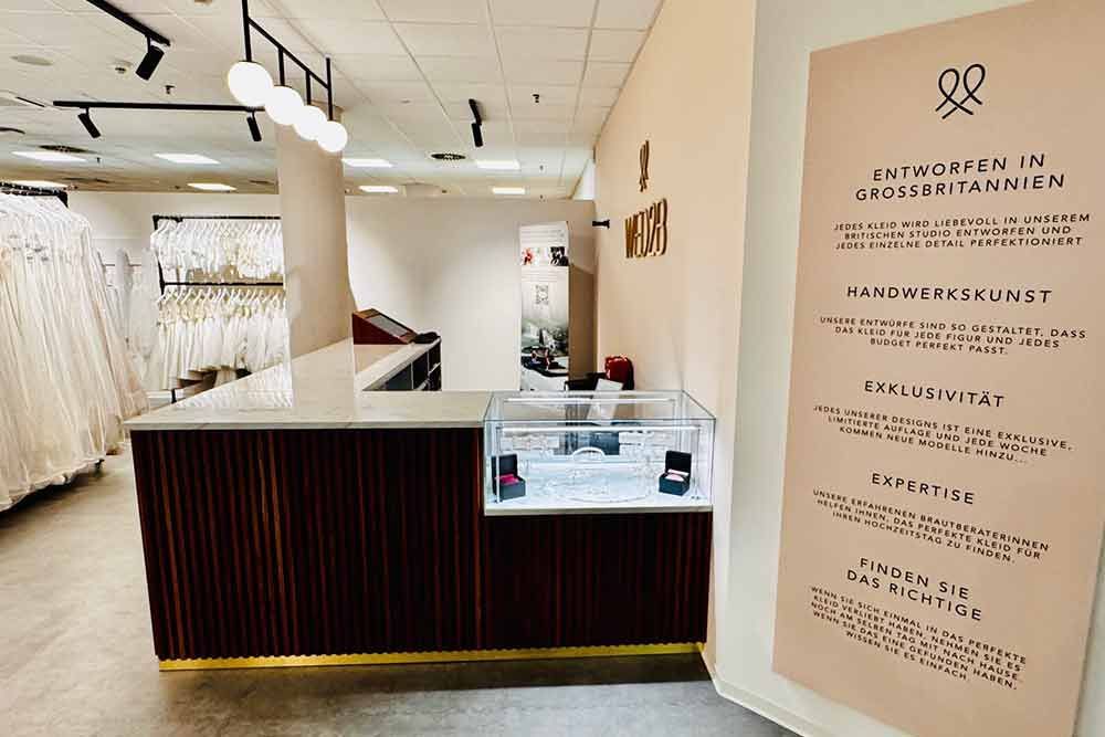 Das Interieur eines Brautgeschäfts mit Empfangstresen, Vitrine und Kleidern. Ein Plakat listet die angebotenen Dienstleistungen auf.