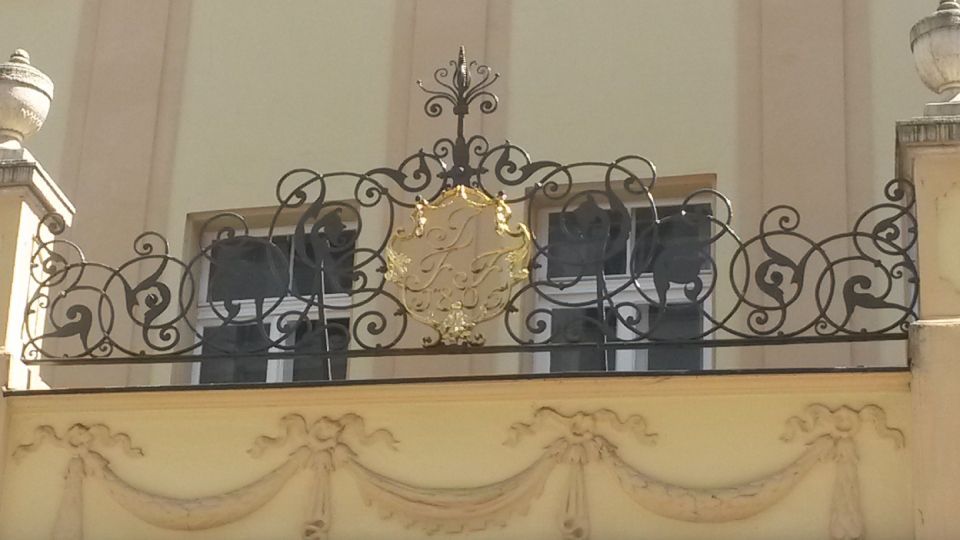 Aufwendig verziertes schmiedeeisernes Geländer mit goldenem Emblem an einer Gebäudefassade. Beige-goldene Farbgebung.