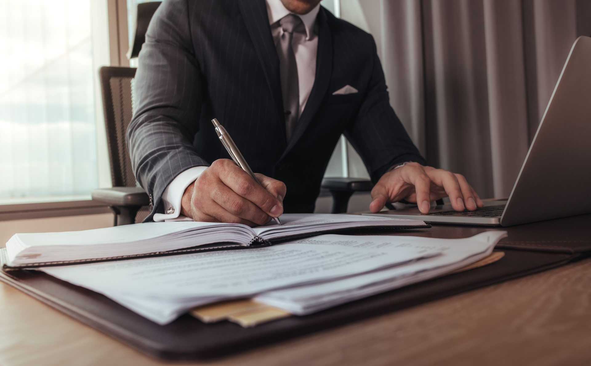 Une personne écrit à son bureau avec un ordinateur portable et des documents.