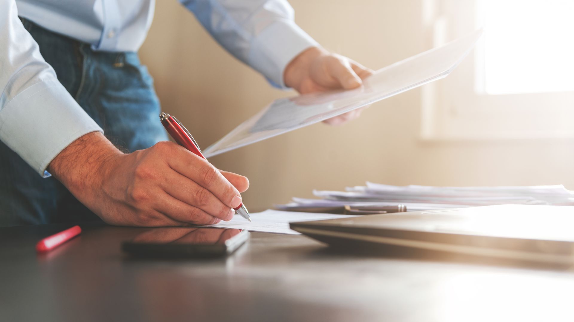 Une personne signe un document, d'autres papiers et un ordinateur portable se trouvent sur le bureau.