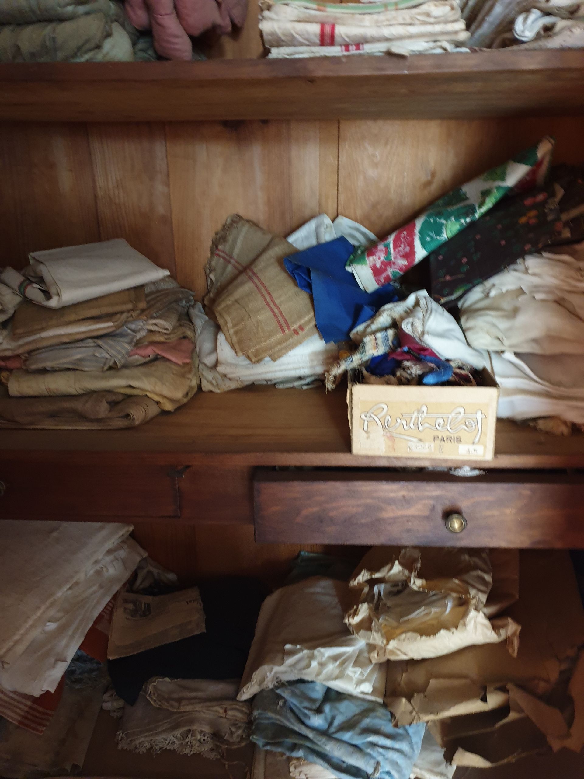 Un meuble en bois rempli de textiles : du linge plié, des tissus à motifs et une petite boîte sur une étagère