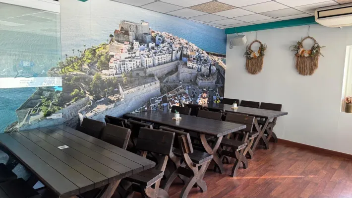 Interior de restaurante con mesas y sillas de madera oscura, un mural de un pueblo costero y tapices decorativos.