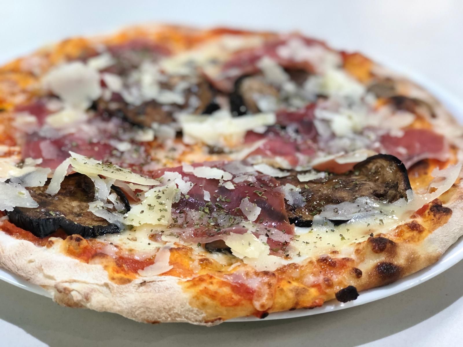 Pizza cubierta con prosciutto, berenjena y queso parmesano en un plato blanco.