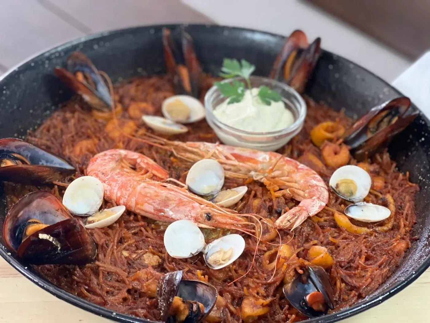 Fideuá de mariscos en sartén, cubierta con camarones, mejillones, almejas y alioli.