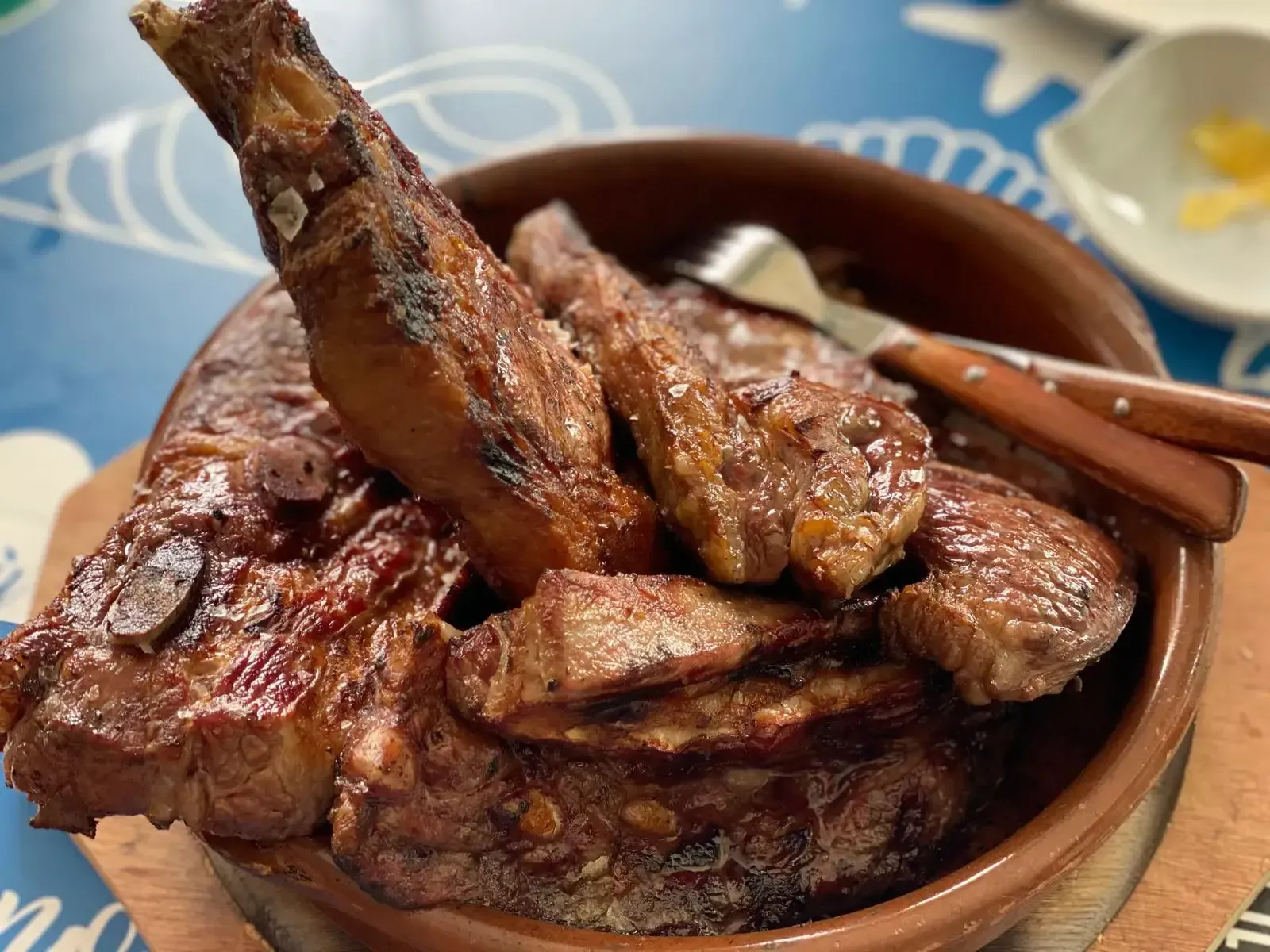 Costillas de cordero a la parrilla en un recipiente marrón con tenedor y cuchillo, sobre una tabla de madera.