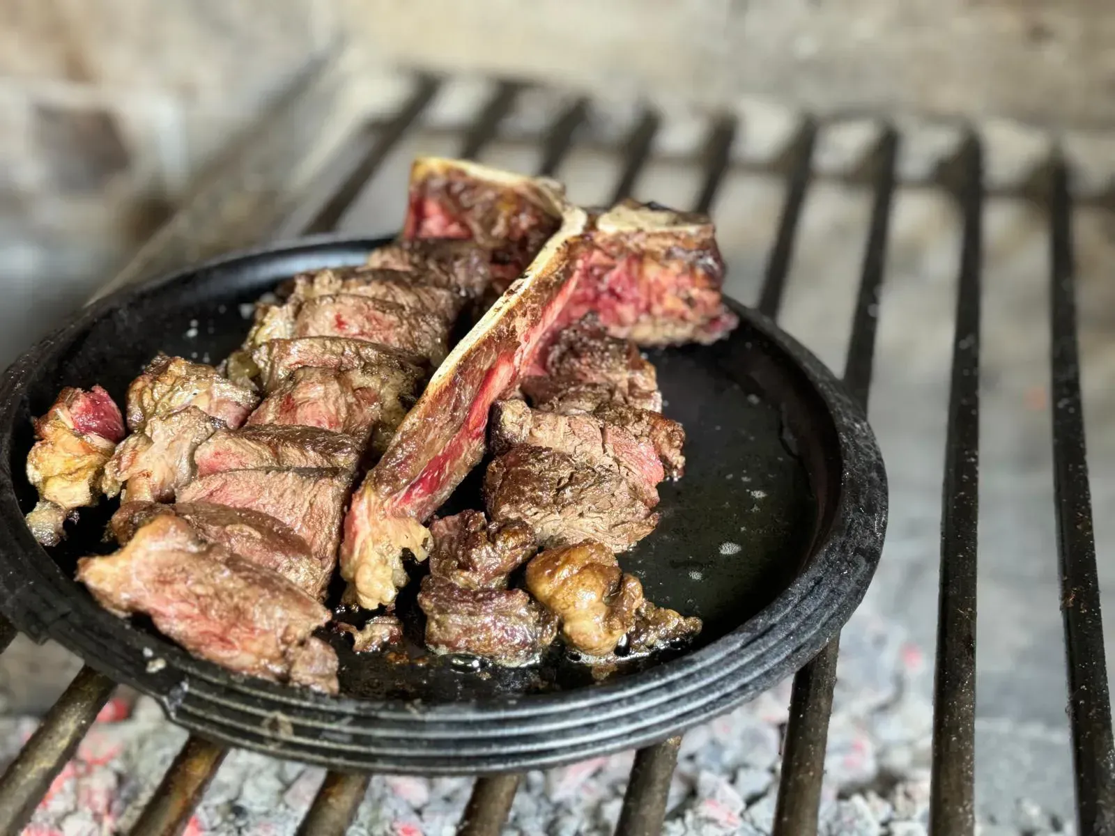 Filete T-bone cortado en rodajas sobre una fuente de hierro fundido negro, apoyado sobre una parrilla de carbón.