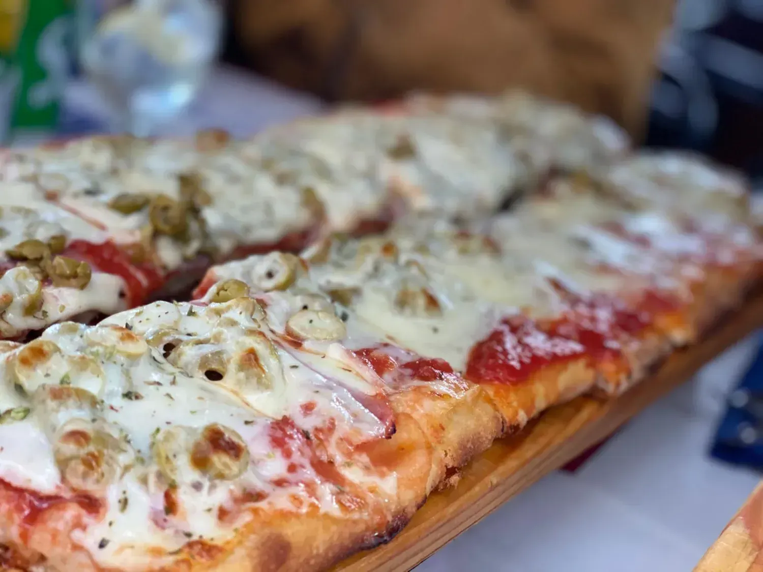 Pizza rectangular cubierta con queso, aceitunas y salsa de tomate, presentada en una bandeja de madera.