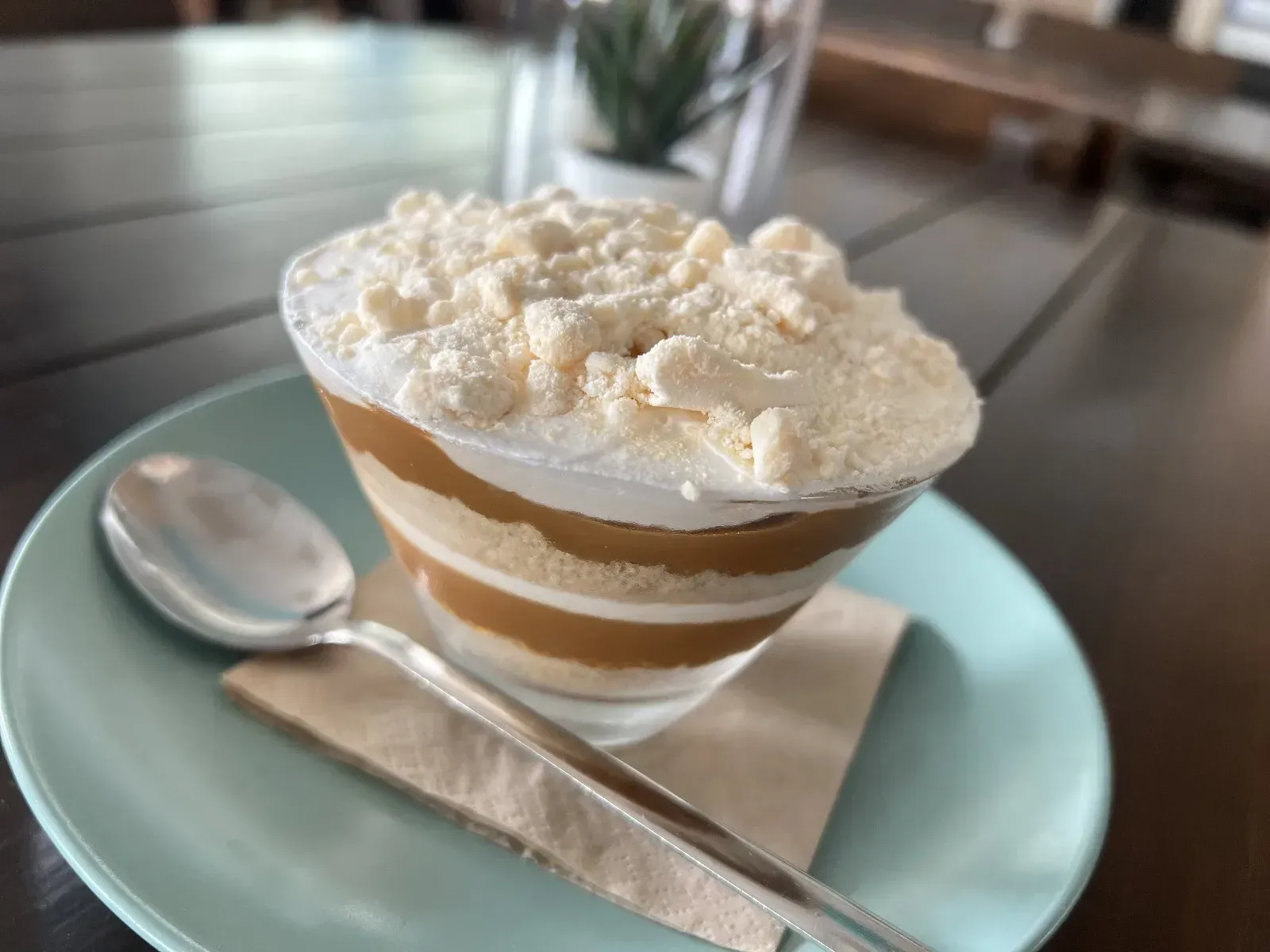 Parfait de postre en un recipiente de vidrio sobre un plato azul, con una cuchara y una servilleta, sobre una mesa de madera.