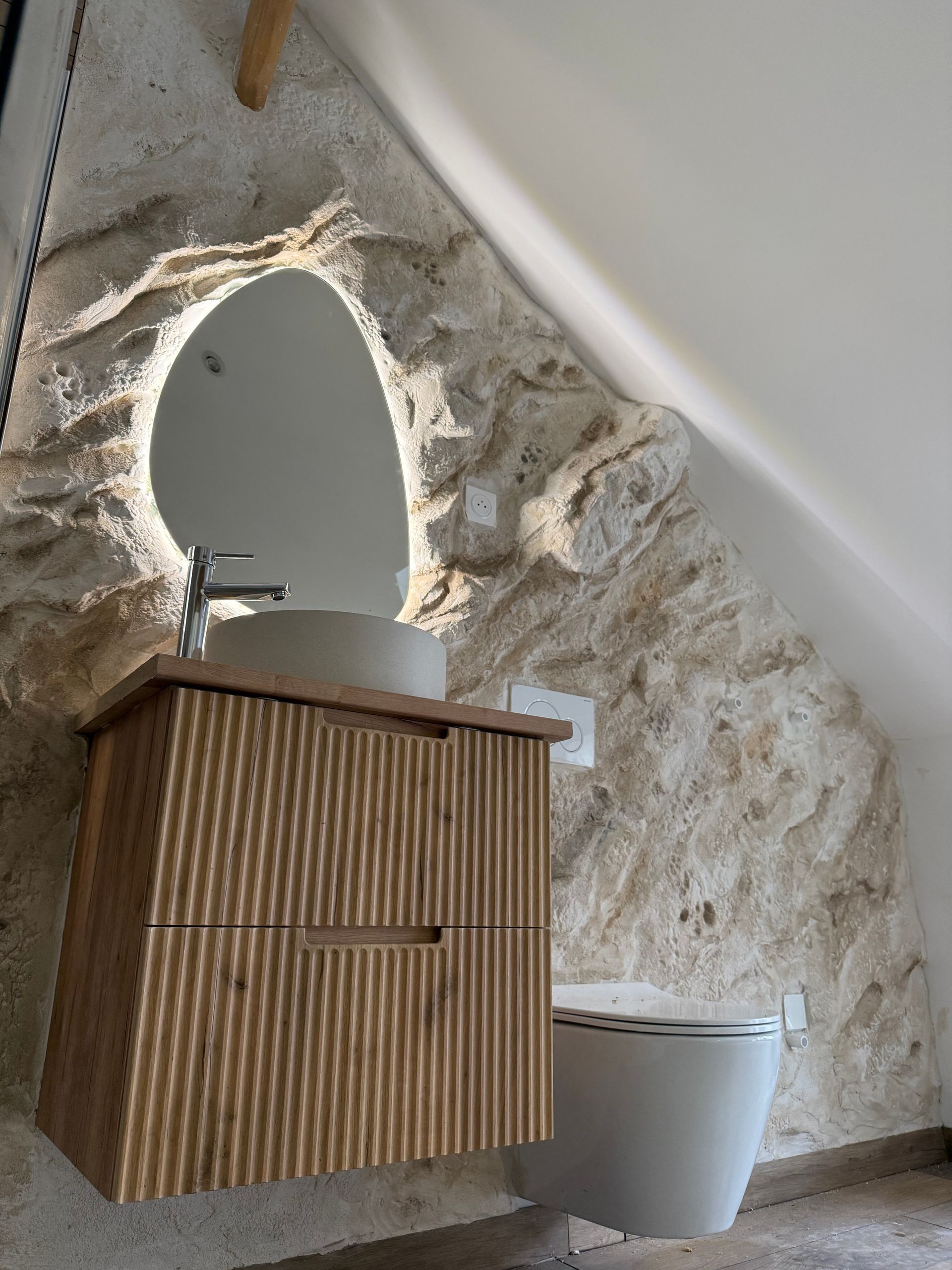 Salle de bains avec mur en pierre apparente, meuble-lavabo en bois, miroir ovale et toilettes.