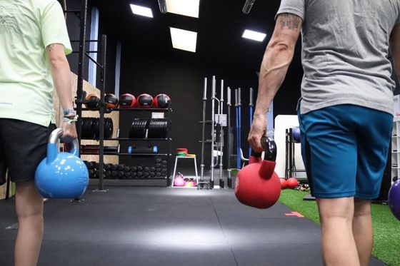 Dos personas en un gimnasio con pesas rusas, listas para hacer ejercicio. Una pesa azul y otra roja.
