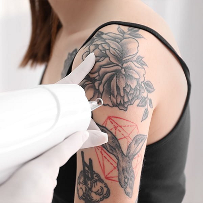 Der Arm einer Person wird tätowiert. Eine weiß behandschuhte Hand hält eine Tätowiermaschine mit einem floralen Tattoo und geometrischem Muster auf dem Arm.