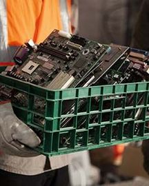 Caisse en plastique contenant plusieurs cartes électroniques et composants informatiques usagés, transportée par une personne équipée de gants de manutention.