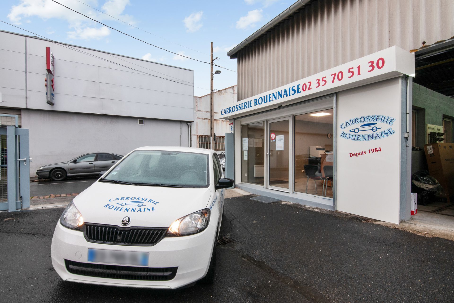 Une voiture blanche arborant le logo de la Carrosserie Rouennaise garée devant leur atelier.