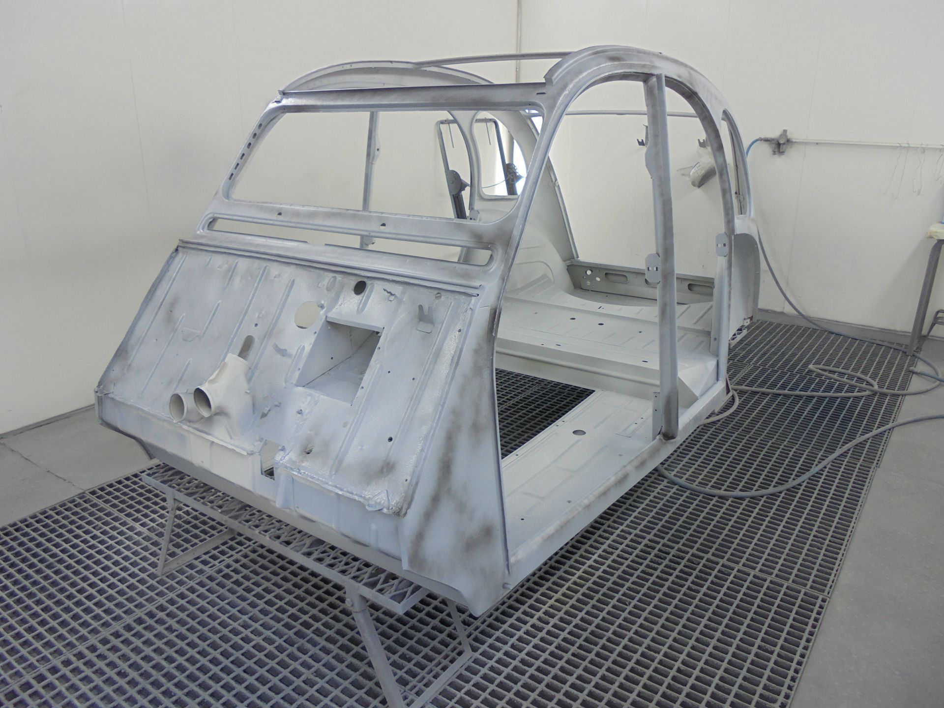 Le châssis métallique d'une Citroën 2CV repose dans une cabine de peinture automobile.