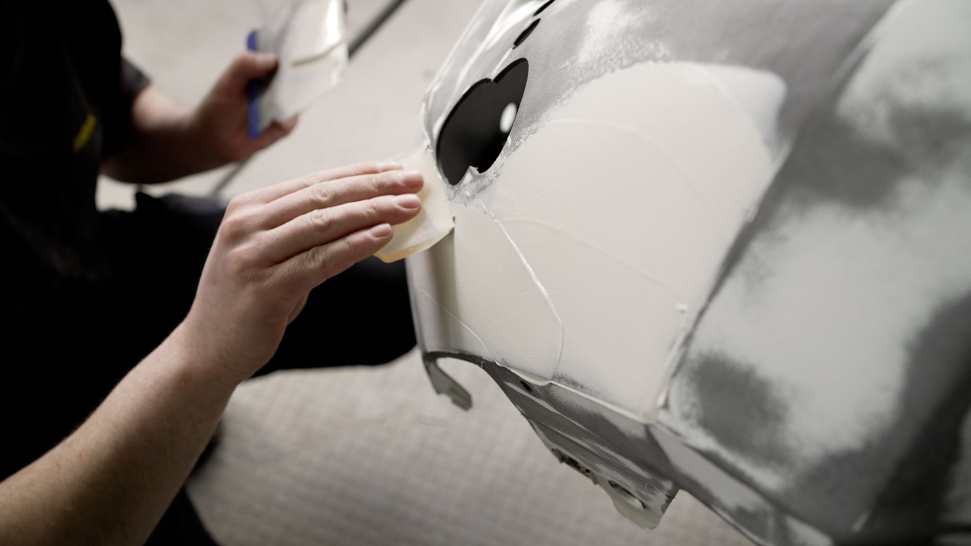 Un ouvrier utilise une spatule en plastique pour appliquer du mastic sur un pare-chocs de voiture.