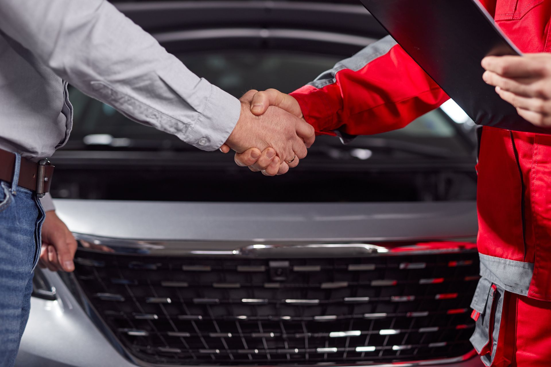 Une personne en tenue décontractée serre la main d'un mécanicien en salopette rouge devant une voiture.