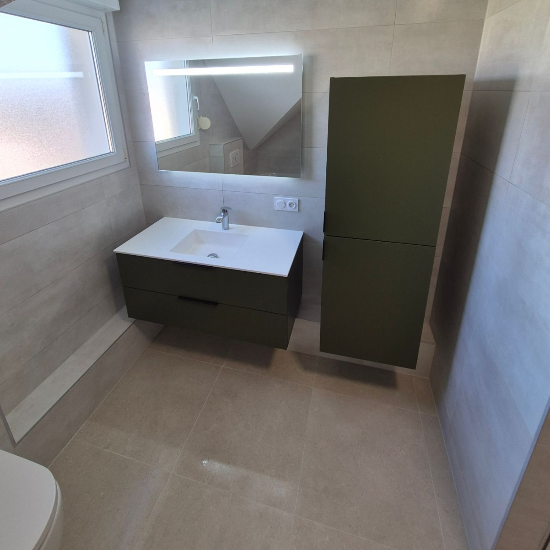 Salle de bains avec meuble-lavabo et armoire vert olive, lavabo blanc, grand miroir et fenêtre.