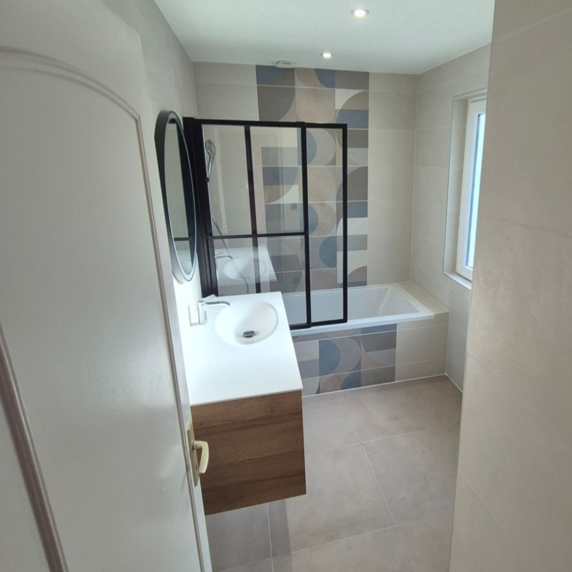 Salle de bains avec lavabo blanc, baignoire et carrelage mural à motifs. Meubles marron et paroi de douche noire.