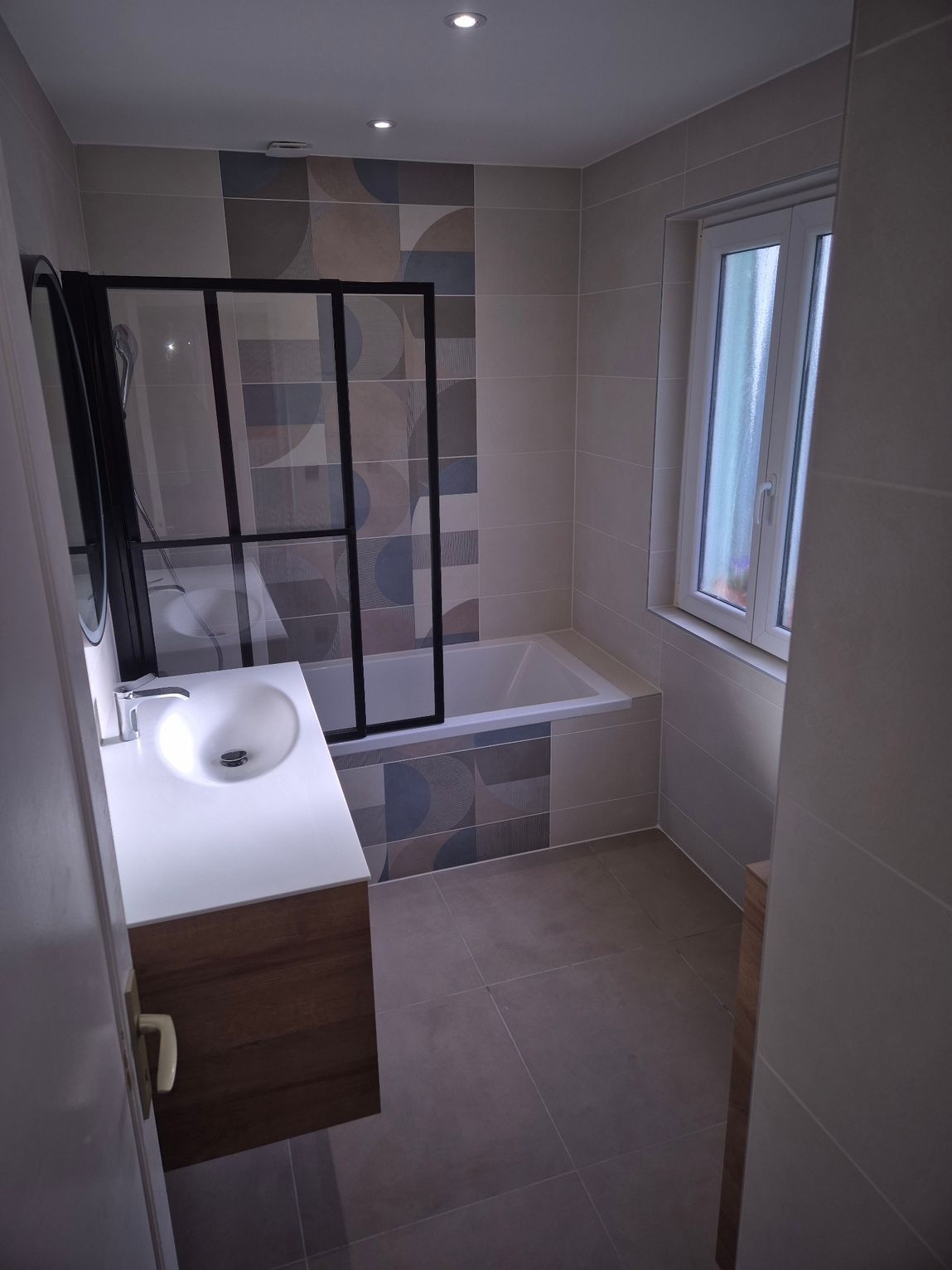 Salle de bains avec lavabo blanc, meuble en bois, baignoire, paroi de douche en verre et fenêtre.