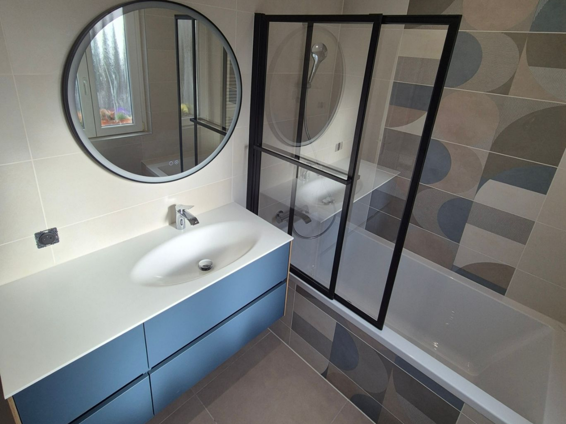 Salle de bains moderne avec meuble-lavabo bleu, miroir rond et carrelage à motifs. Douche à cadre noir au-dessus de la baignoire.