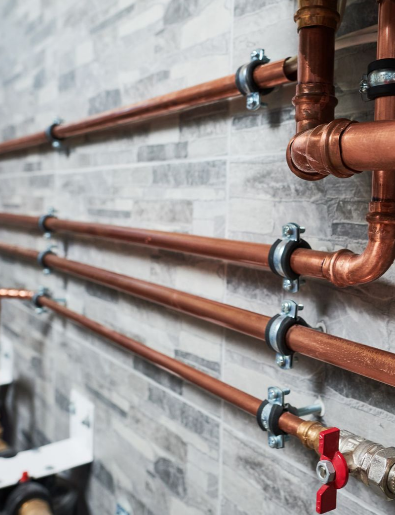 Tuyaux de cuivre fixés à un mur carrelé gris, avec une vanne rouge visible.