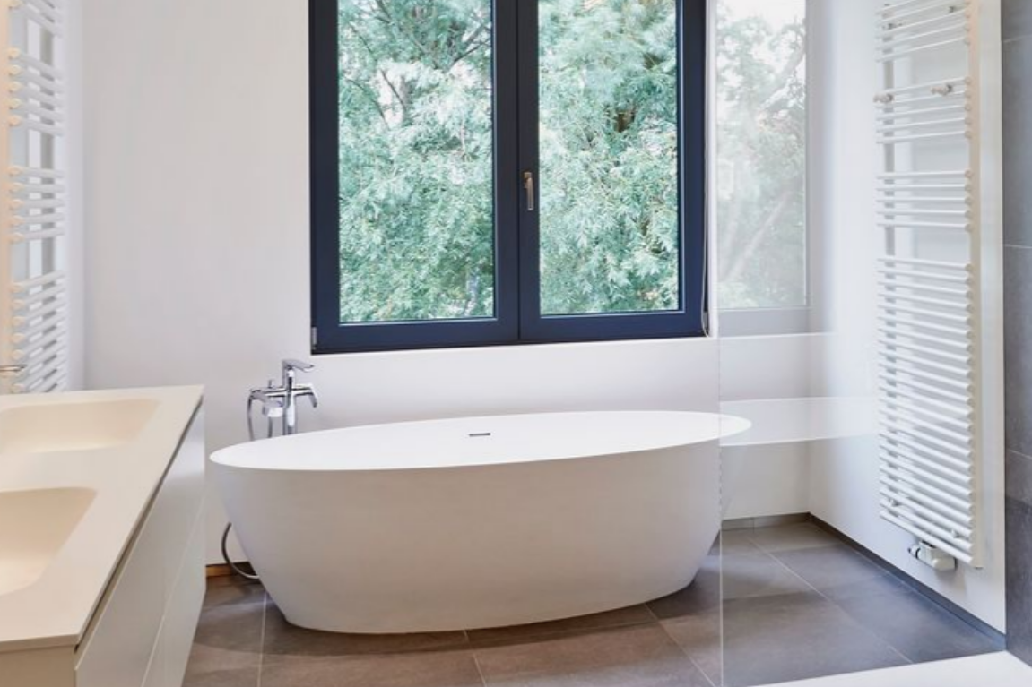 Salle de bains moderne blanche avec baignoire ovale près d'une fenêtre ; sol carrelé gris, murs blancs.