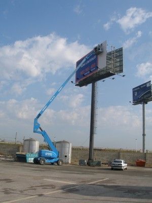 Una grúa azul está levantando una gran valla publicitaria en un estacionamiento.