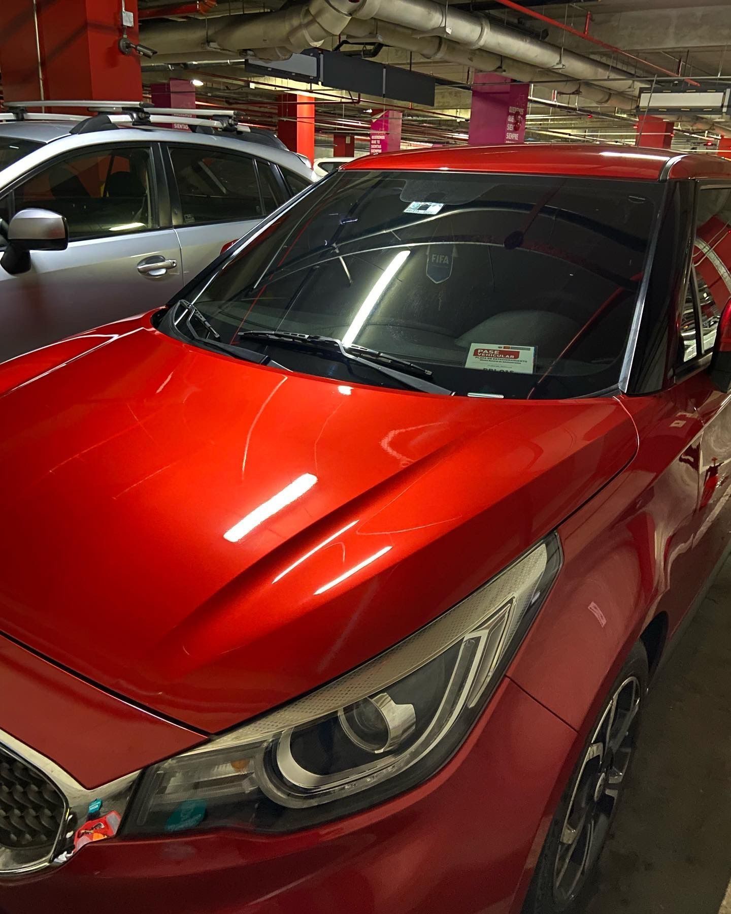 Un coche rojo aparcado en un garaje con el parabrisas roto. Se ve otro coche a su izquierda.