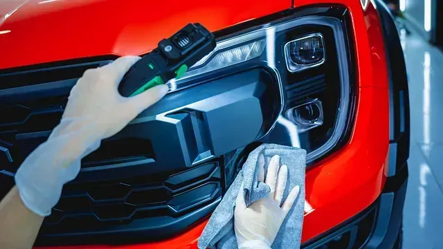 Manos en guantes limpiando un faro rojo de coche con una herramienta y un paño.