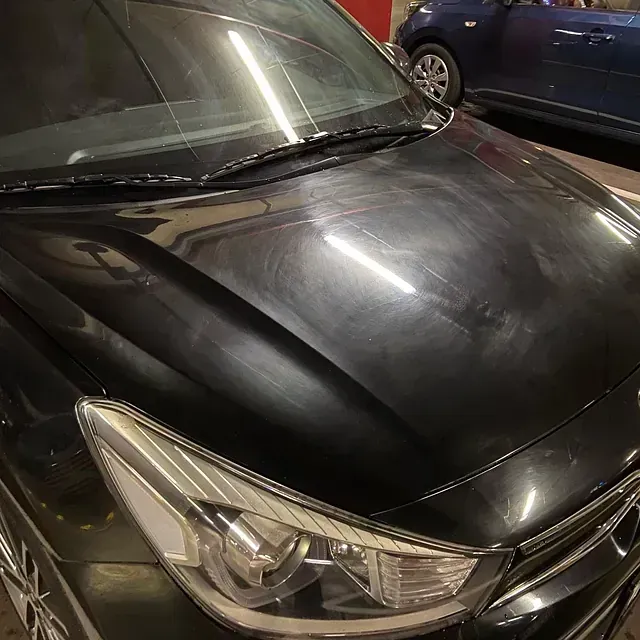 Capó de coche negro con marcas de remolino visibles bajo las luces brillantes del techo, estacionado en el interior.