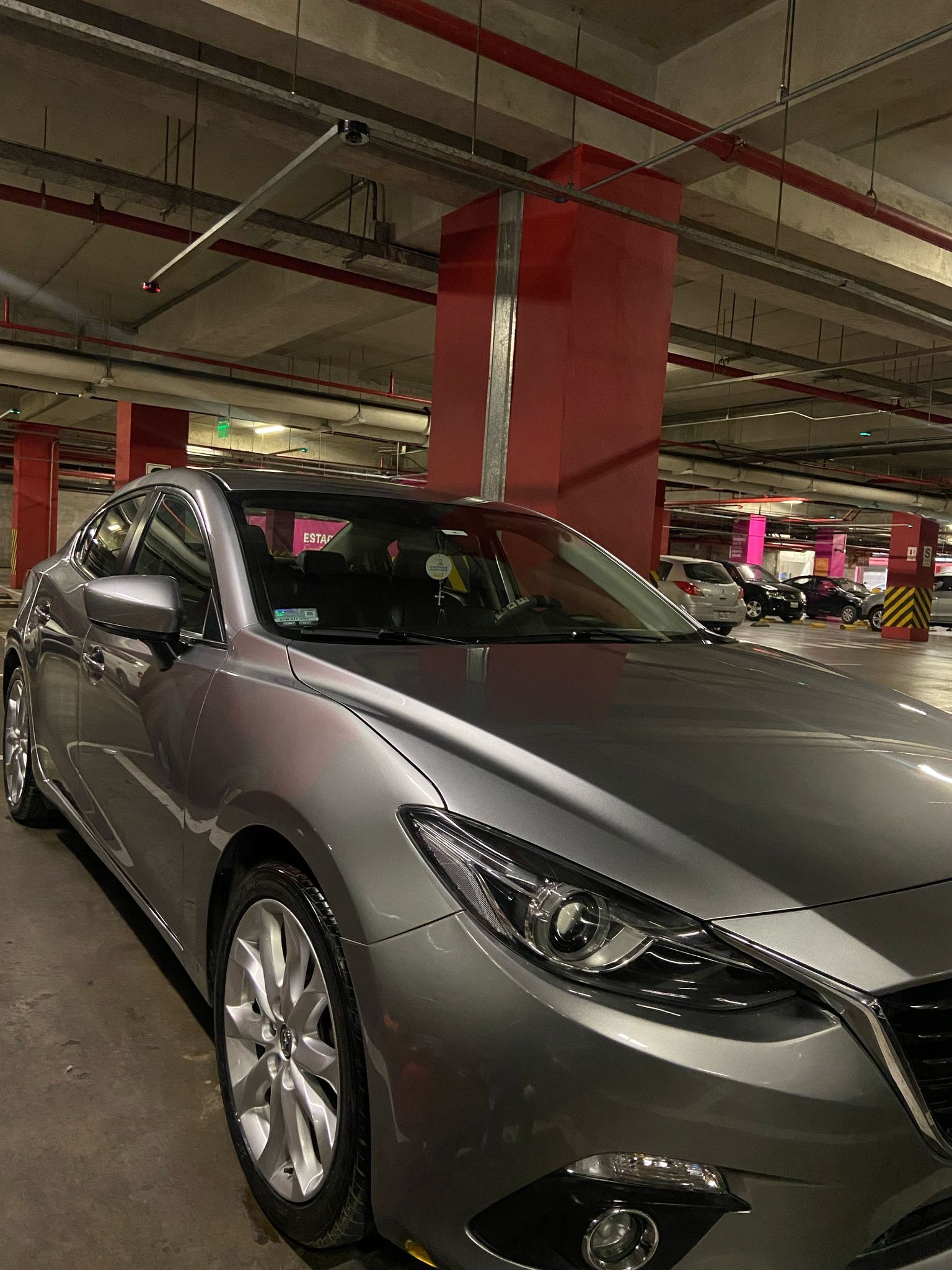 Mazda 3 gris estacionado en un estacionamiento de varios niveles con columnas rojas e iluminación superior.